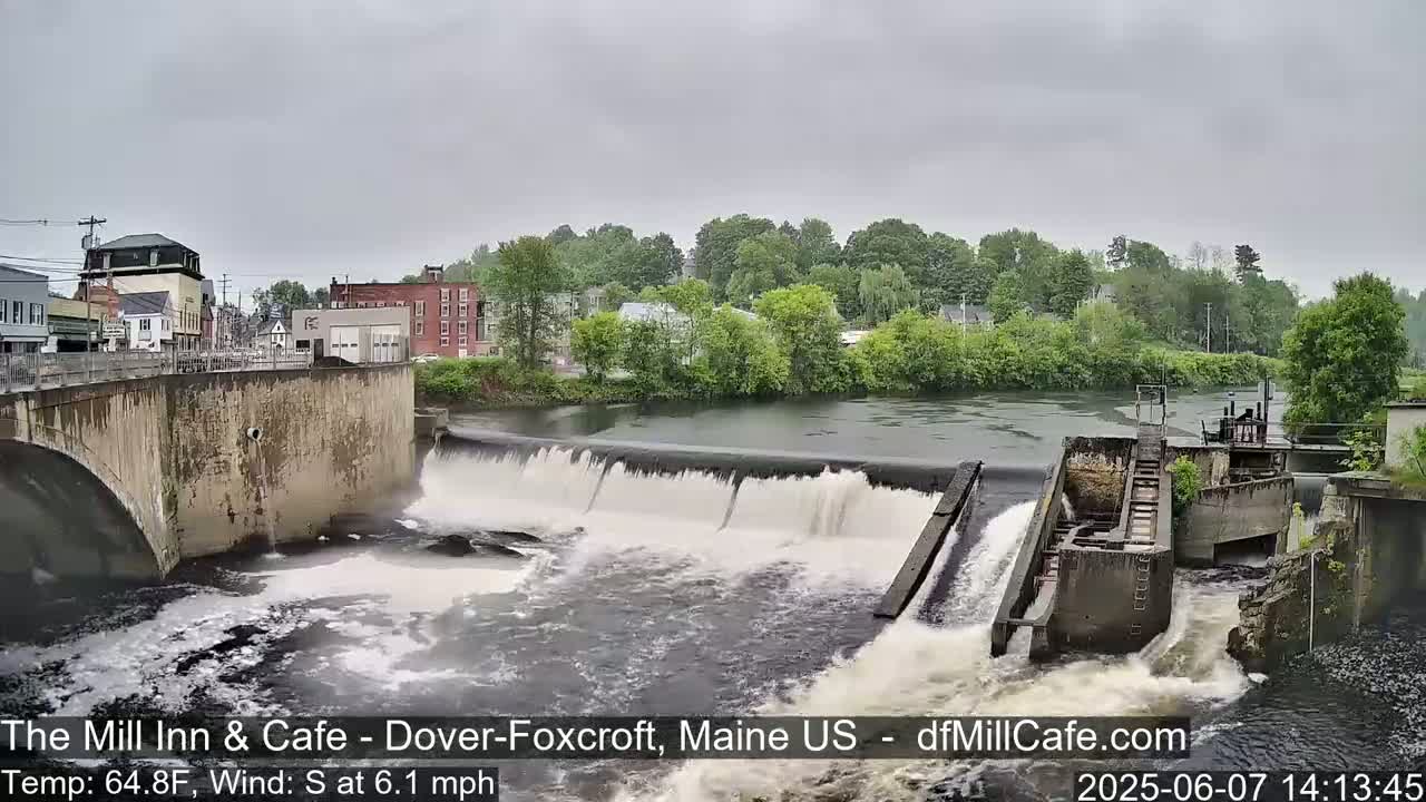 A river flows over a dam in front of a small town under an overcast sky.