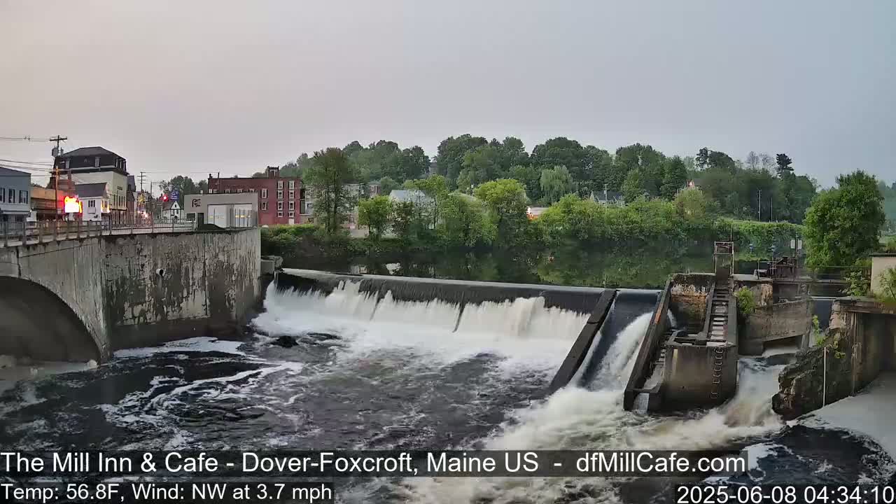 Water flows over a dam in a town under an overcast sky.