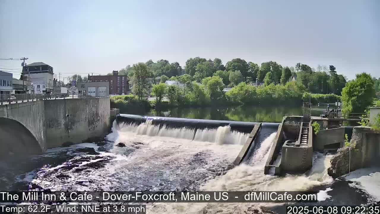 A dam releases water into a churning river, with a town and green trees visible in the background under a clear sky.