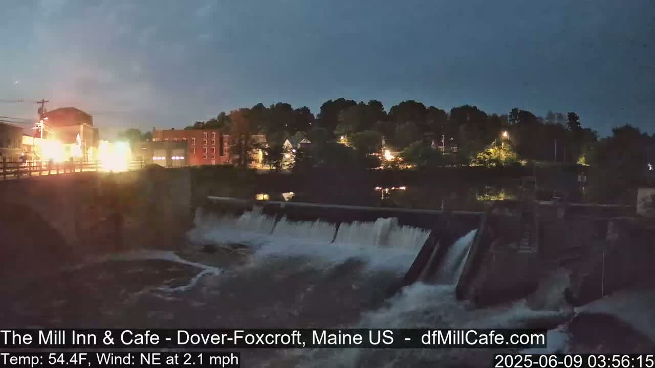 A nighttime view of a dam with water flowing over it, illuminating a town across a river under a twilight sky with a light northeast wind.