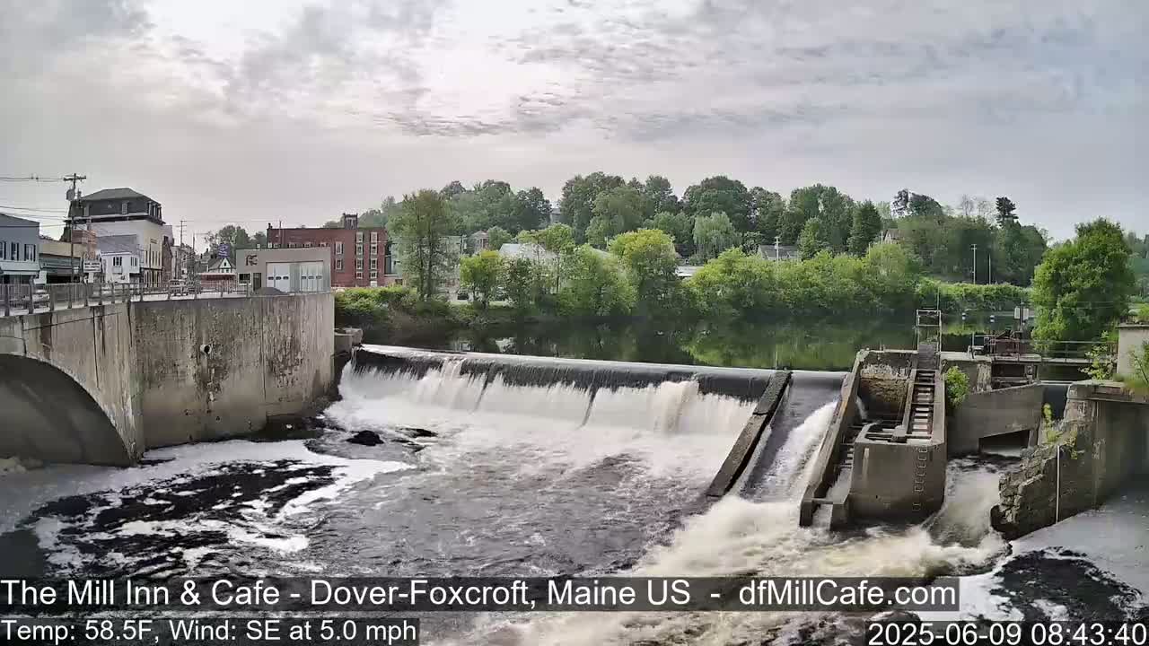 A river flows over a dam in a town, under a mostly cloudy sky.