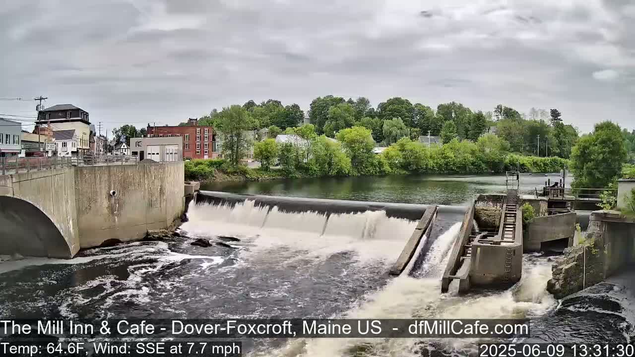 A river flows over a dam in front of a small town under an overcast sky.