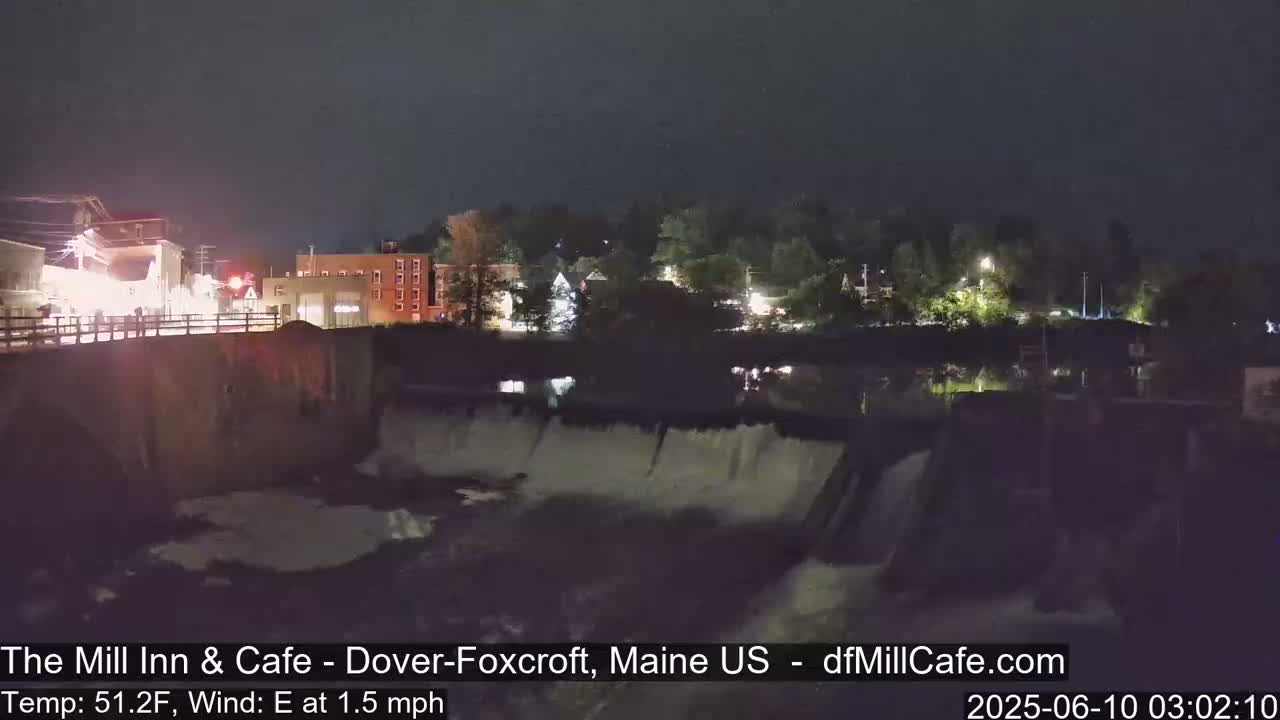 A nighttime view of a town's waterfront, showing a dam with water flowing over it, under a dark, clear sky.