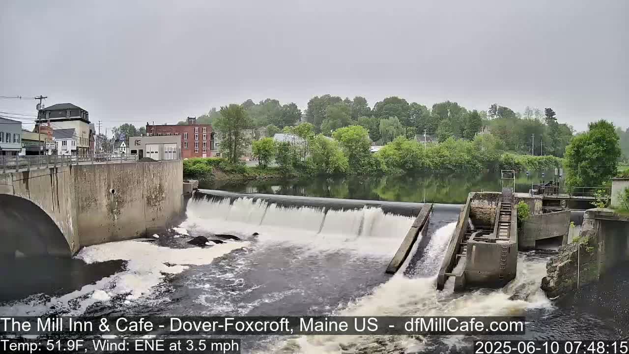 A river flows over a dam in a town under an overcast sky.