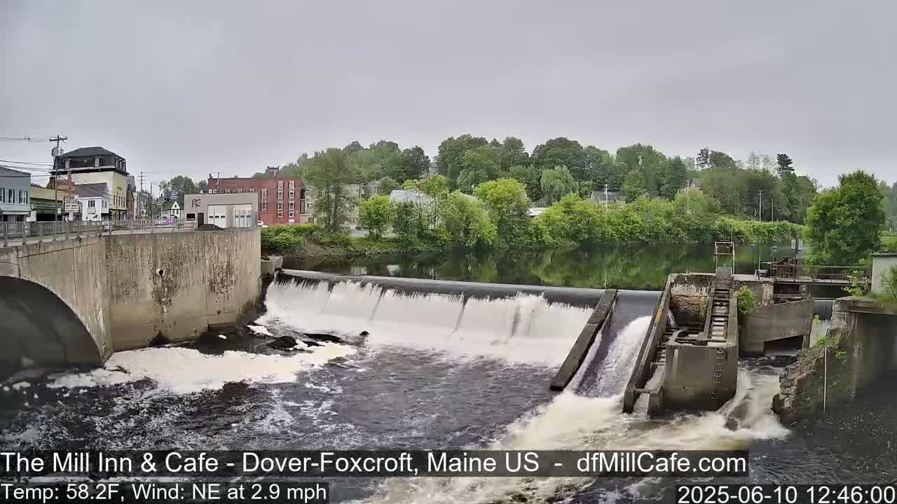 A dam with water flowing over it, situated in front of a town with overcast skies.