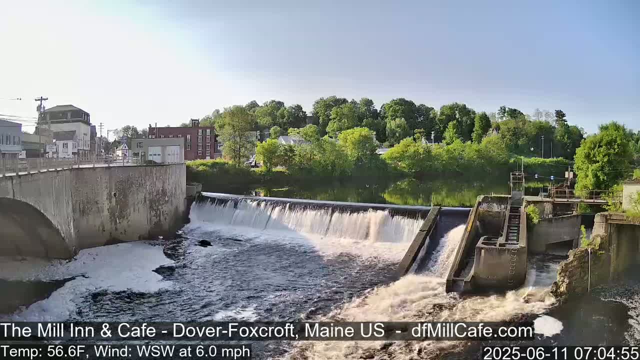 A river flows over a dam in a town, under a clear, bright sky.