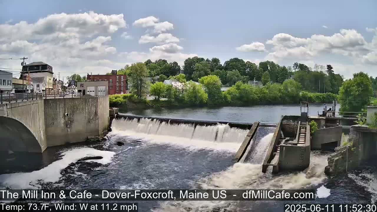 A river flows over a dam, with a town visible in the background under a partly cloudy sky.