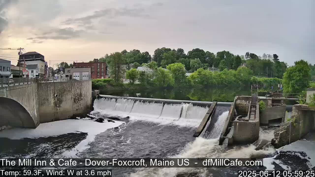 A river flows over a dam in a town under an overcast sky.