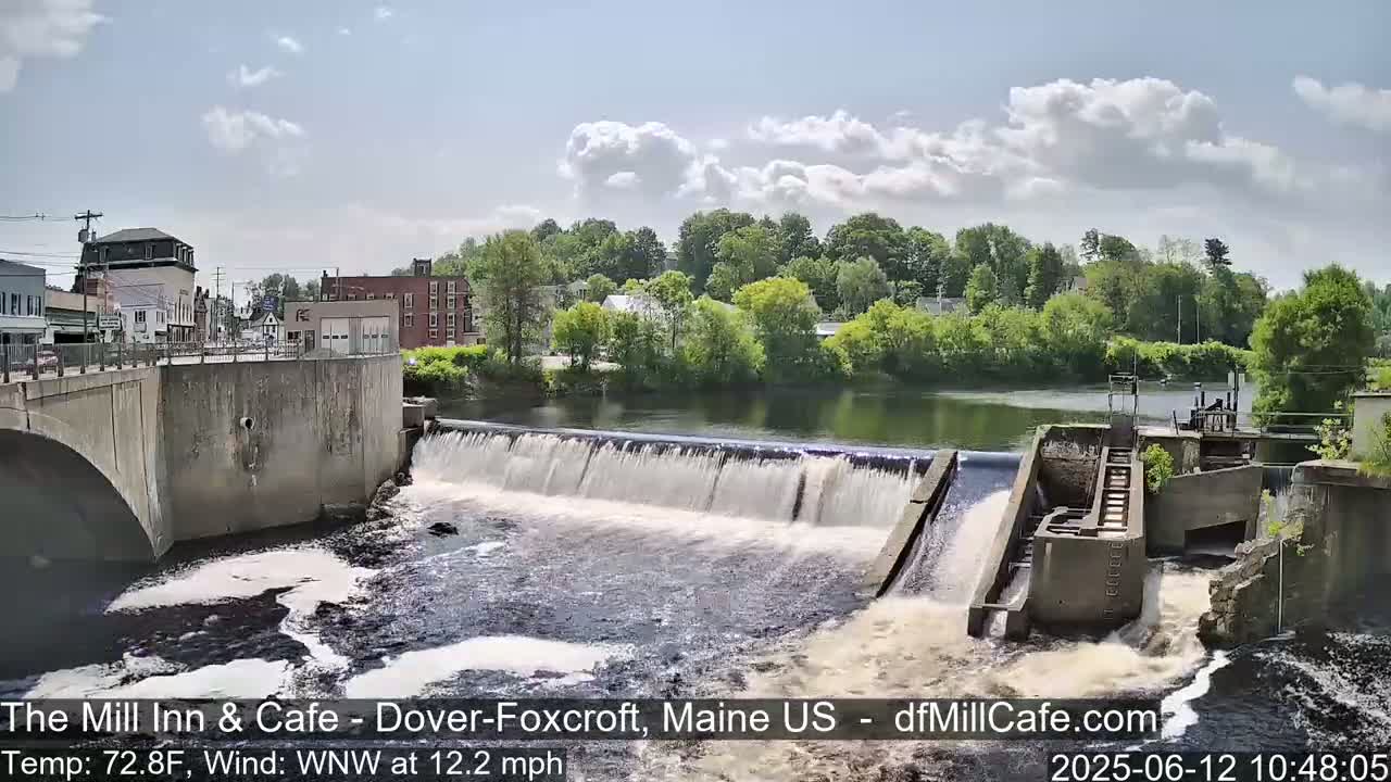 A dam with water flowing over it is situated in front of a town under a partly cloudy sky.