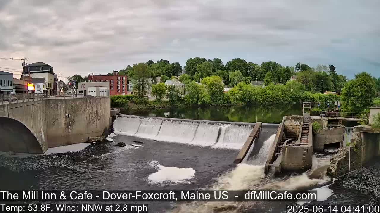 A dam with water flowing over it is situated in front of a town, under a cloudy sky.