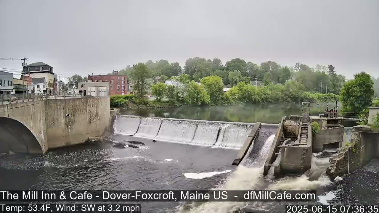 A river flows over a dam beneath a bridge and past buildings on a foggy day.
