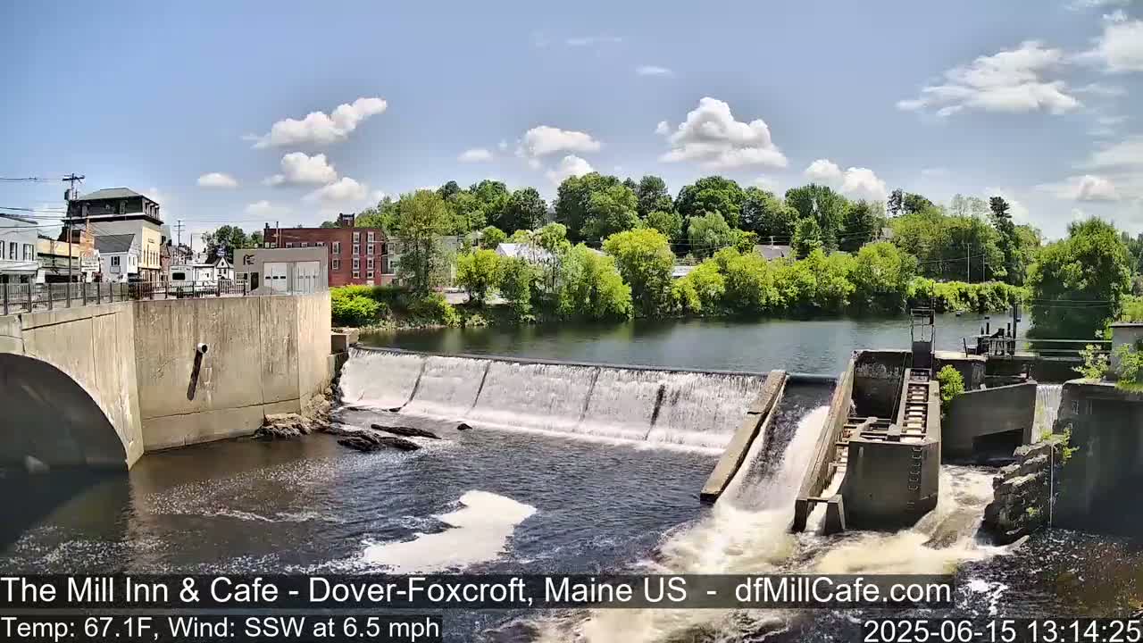 A dam with water flowing over it is situated in a town on a sunny day with partly cloudy skies.