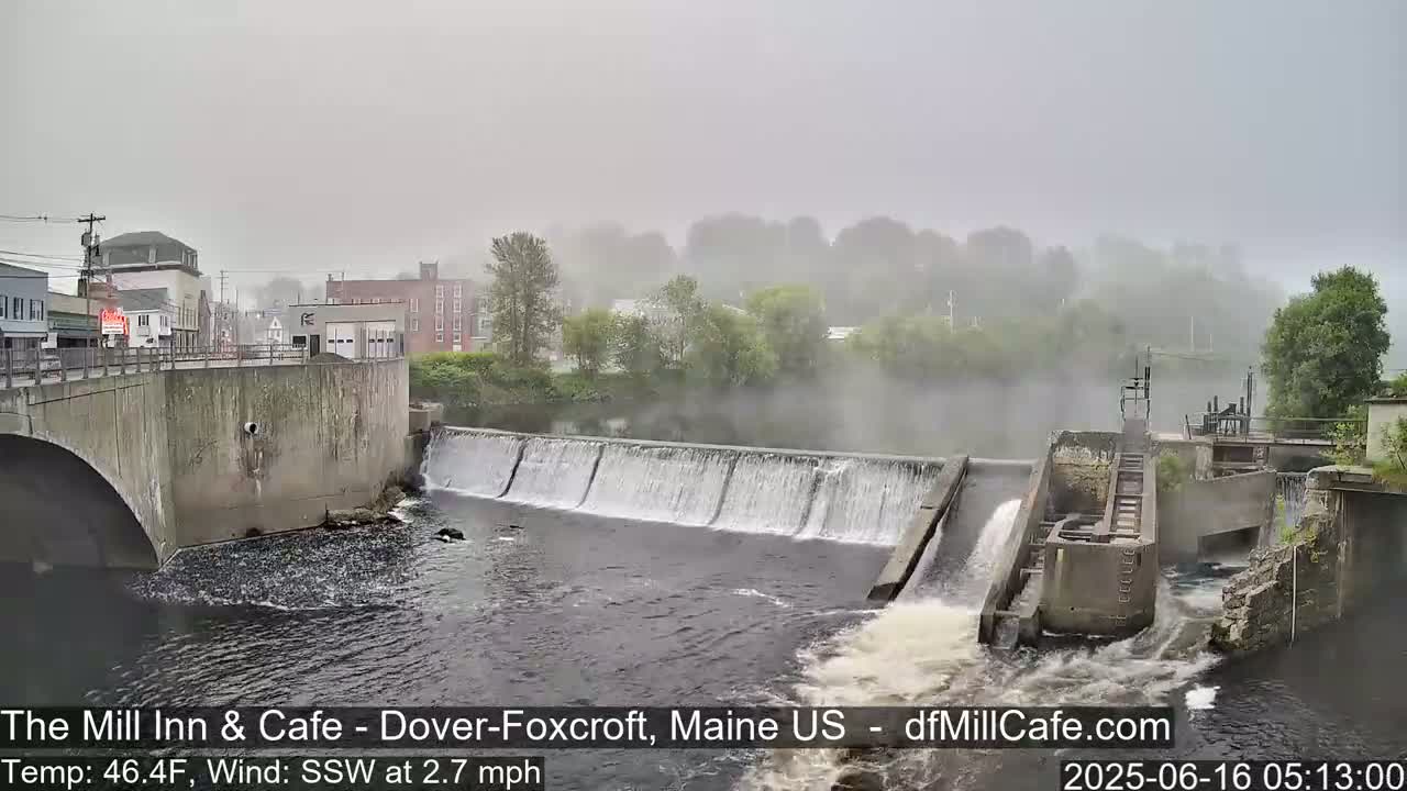 A low-lying dam with water flowing over it is partially obscured by fog, with buildings and trees visible in the background.