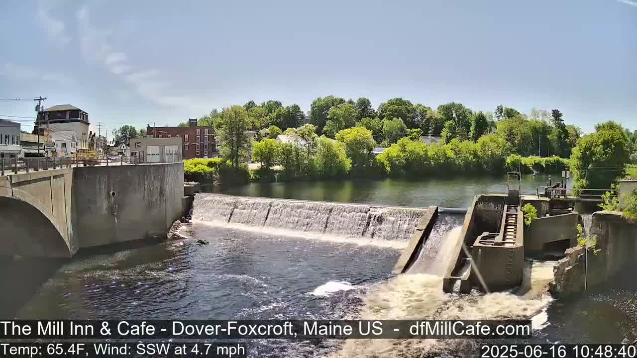 A low dam with water flowing over it into a river, seen from a bridge overlooking a small town on a sunny day.