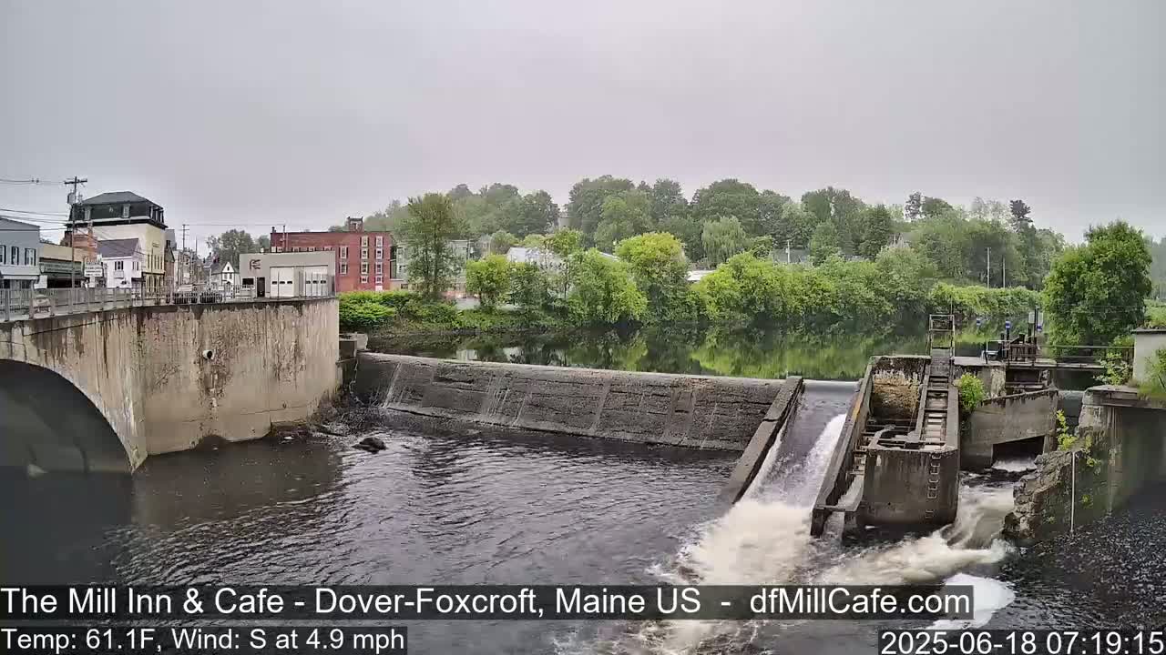 A river flows under a bridge past a dam with water flowing over it, on an overcast day.