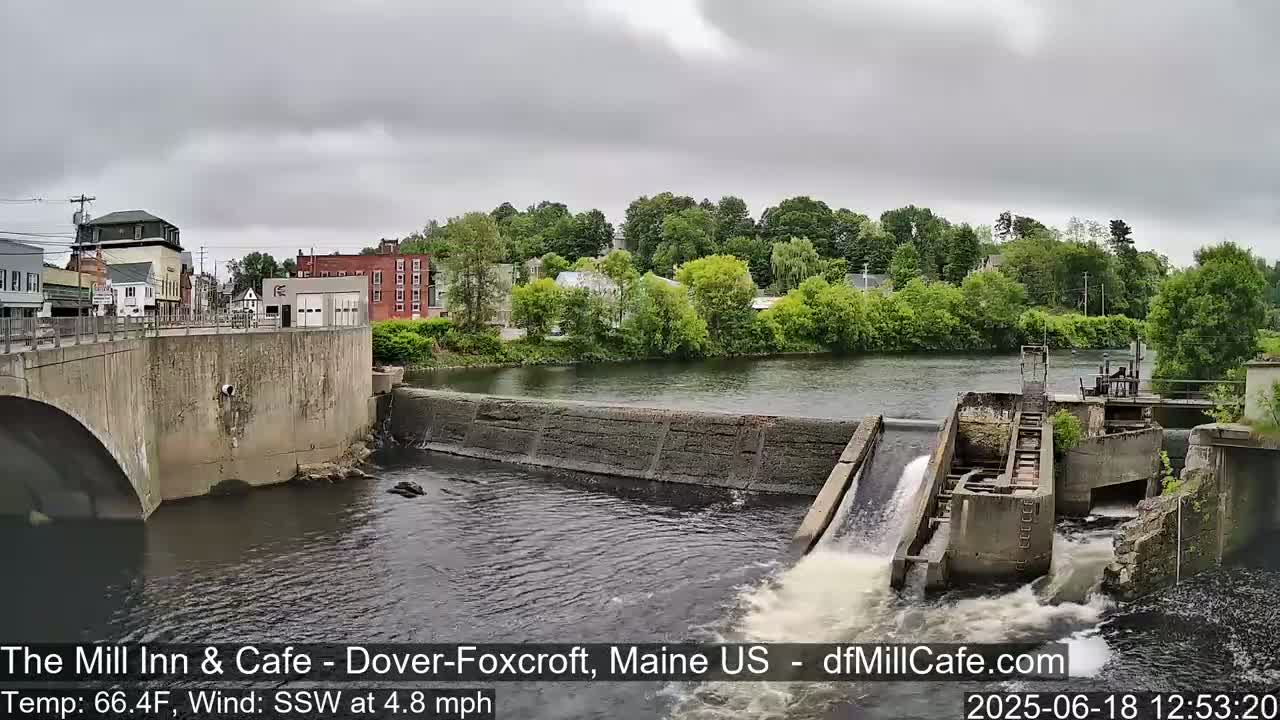 A river flows under a bridge and over a dam, with a town visible in the background under an overcast sky.