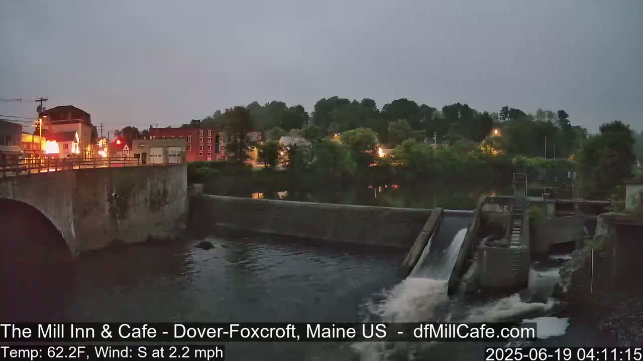 A river flows under a bridge past a town at dusk, with a dam releasing water into the river.