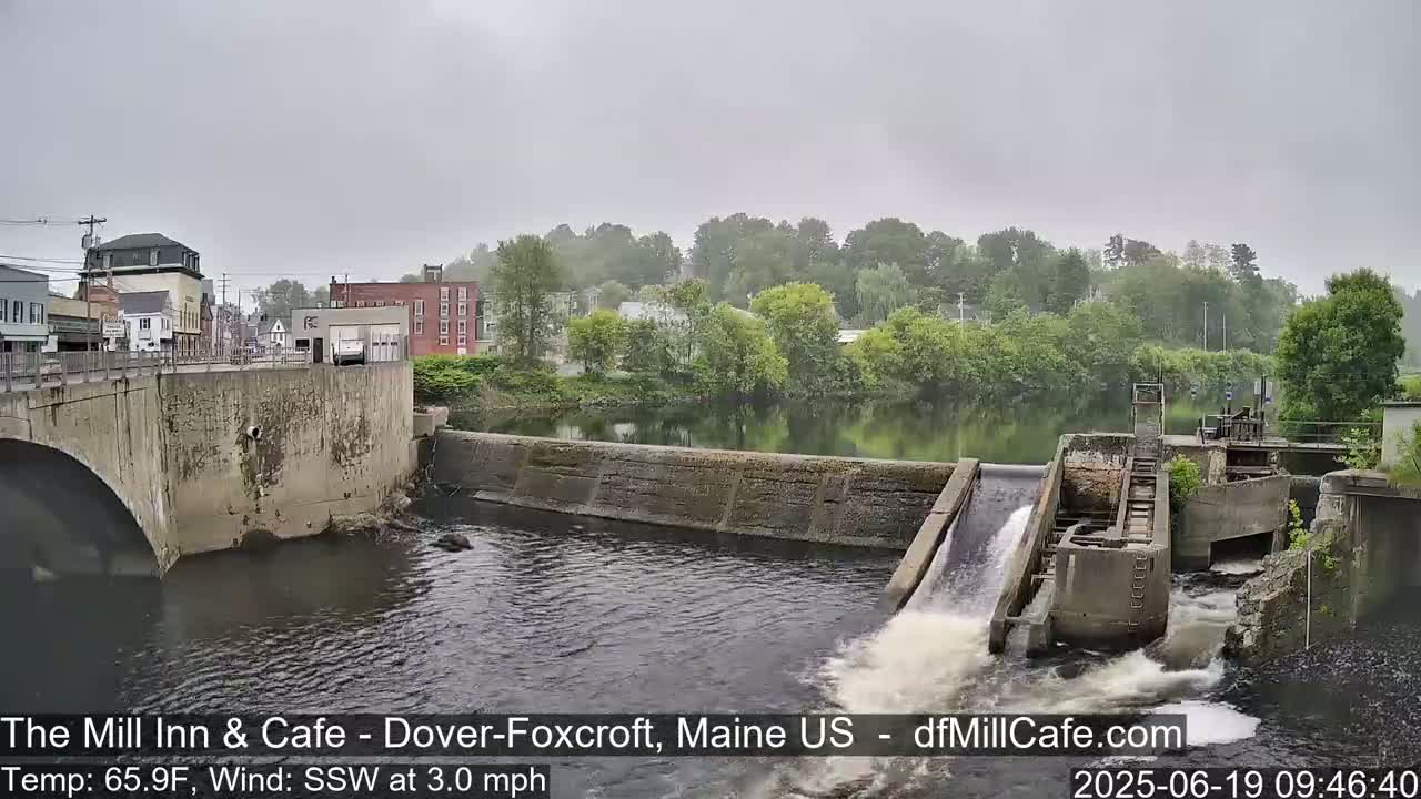 A river flows under a bridge past a dam with water cascading over it, on an overcast day.