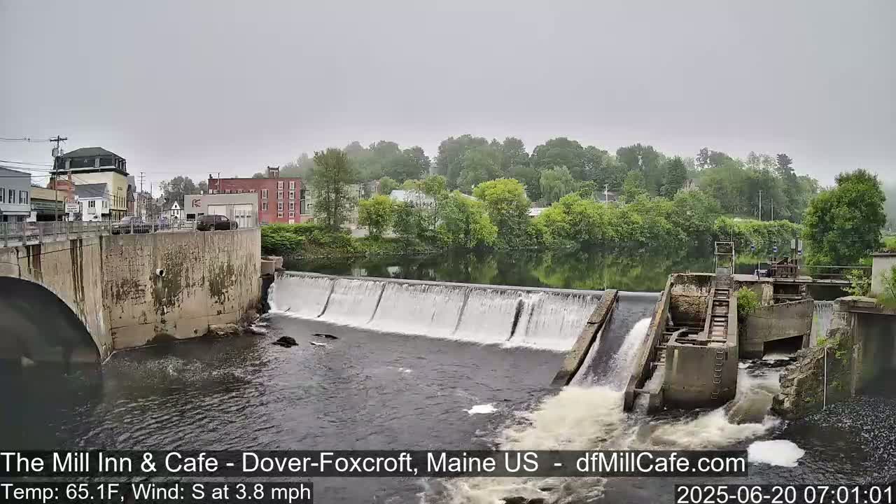 A dam with water flowing over it is situated in front of a town on an overcast day.