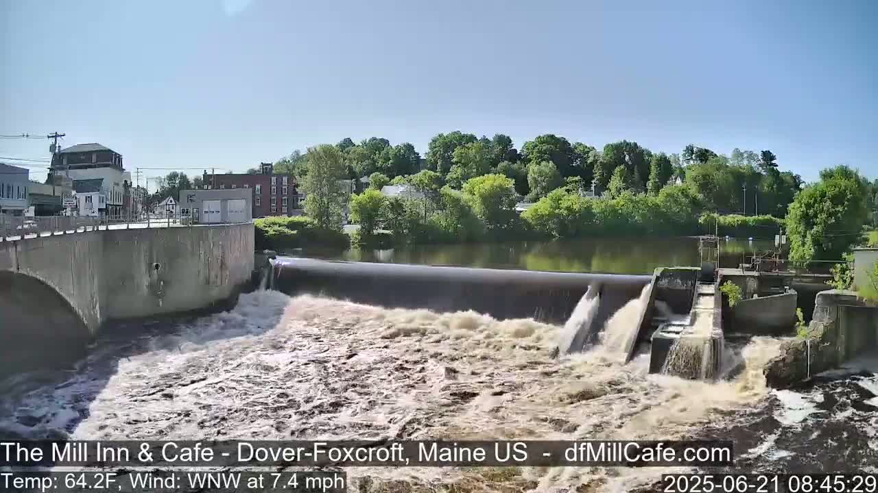 Water rushes over a dam in front of a town under a sunny sky.
