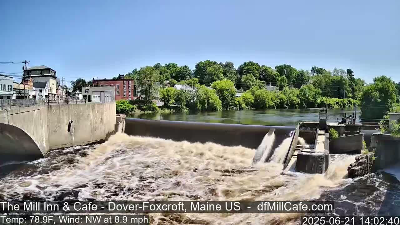 A dam releases water into a churning river on a sunny day, with buildings and trees visible in the background.