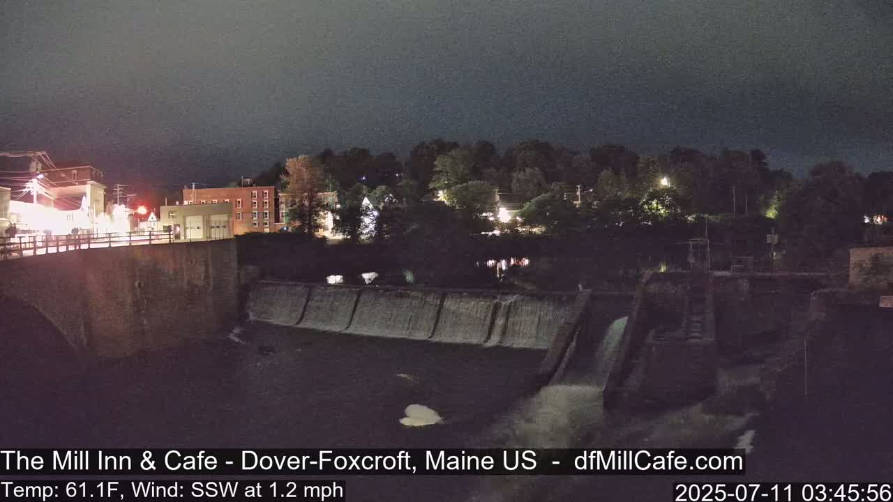 A nighttime view of a dam and river, with buildings and trees illuminated in the background under a dark, overcast sky.