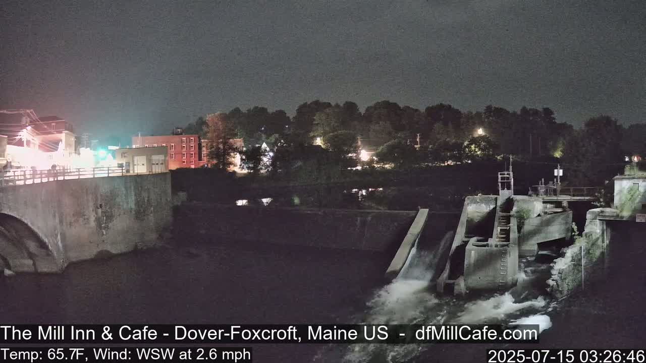 A nighttime view of a dam with water flowing over it, overlooking a town with lights visible under a dark sky.