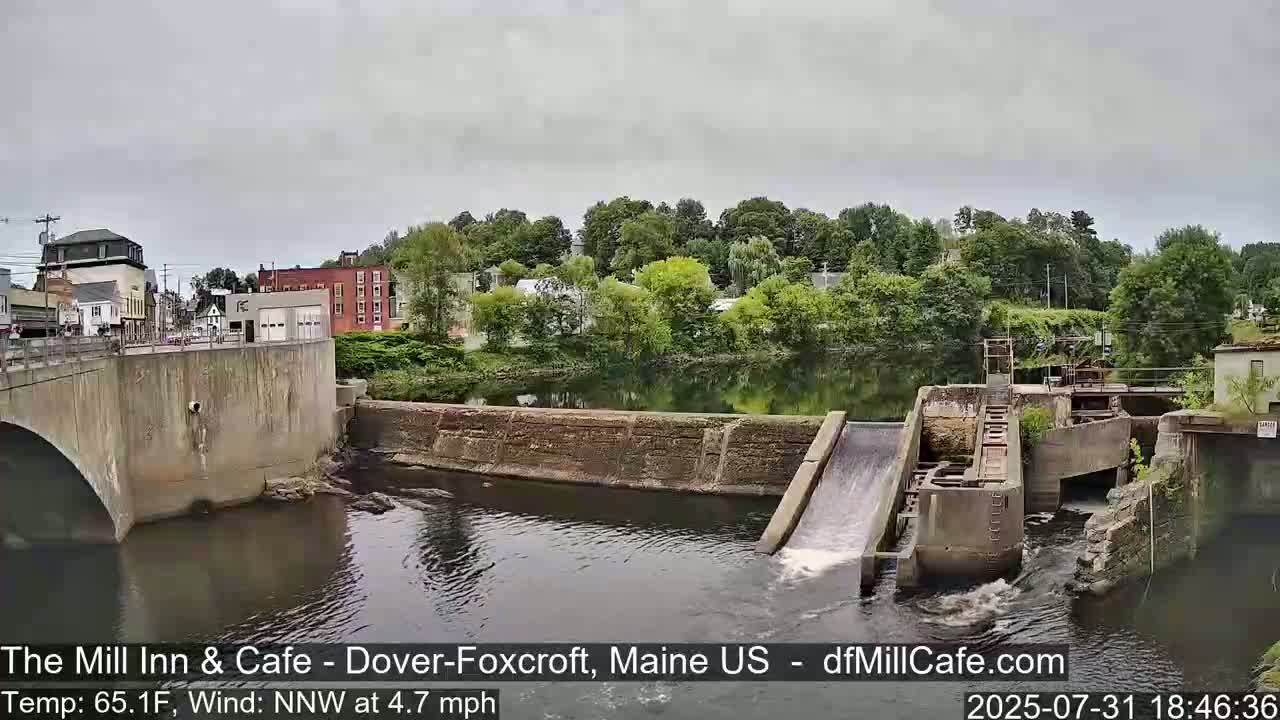 A river flows under a bridge past a dam with water cascading down a spillway, surrounded by green trees and buildings under an overcast sky.