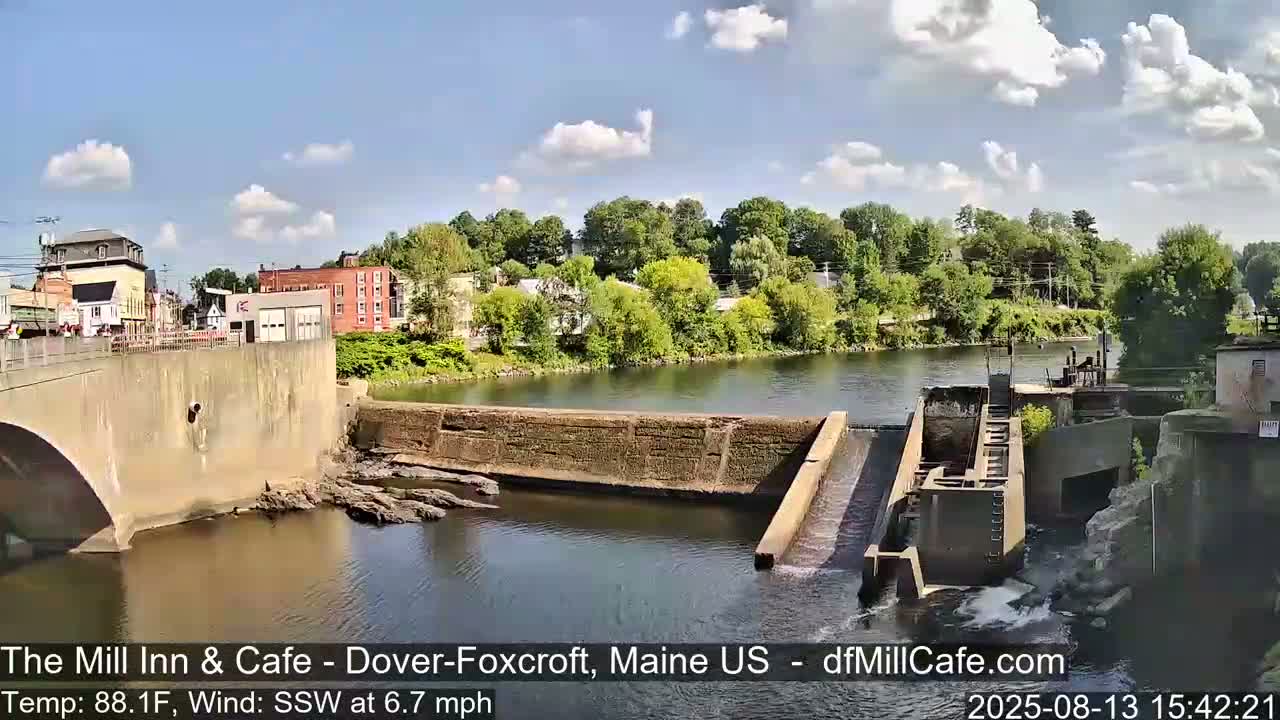 A partly cloudy, sunny day shows a river with a dam and a town visible in the background.