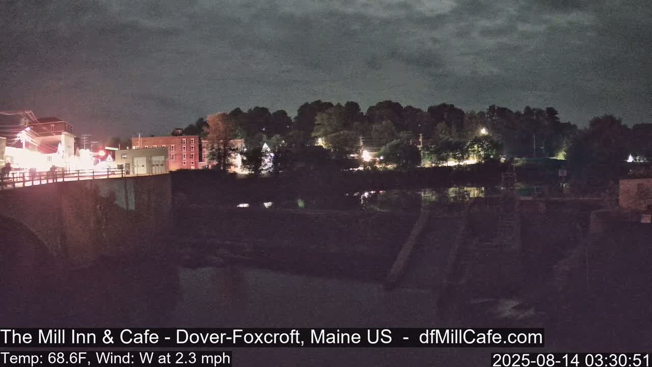 A nighttime view of a town across a river, under a dark, cloudy sky, with a gentle west wind.