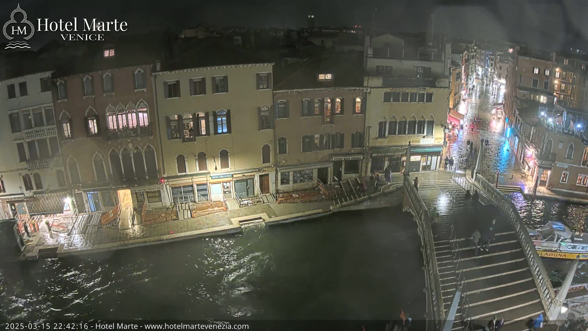 A nighttime view of a Venetian canal shows several buildings along the waterway, a bridge with people walking on it, and a boat on the water, with rain making the surfaces wet.