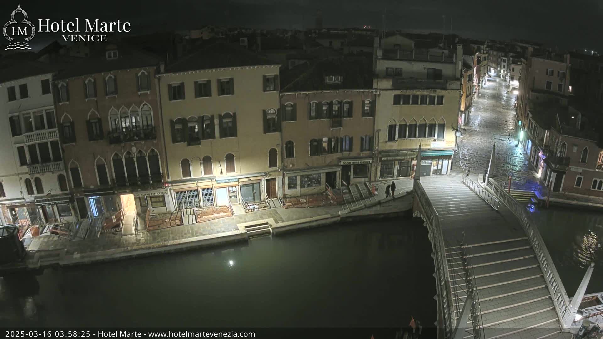 A nighttime, wet view of a canal in Venice, showing several buildings, a bridge with stairs, and a few people walking.