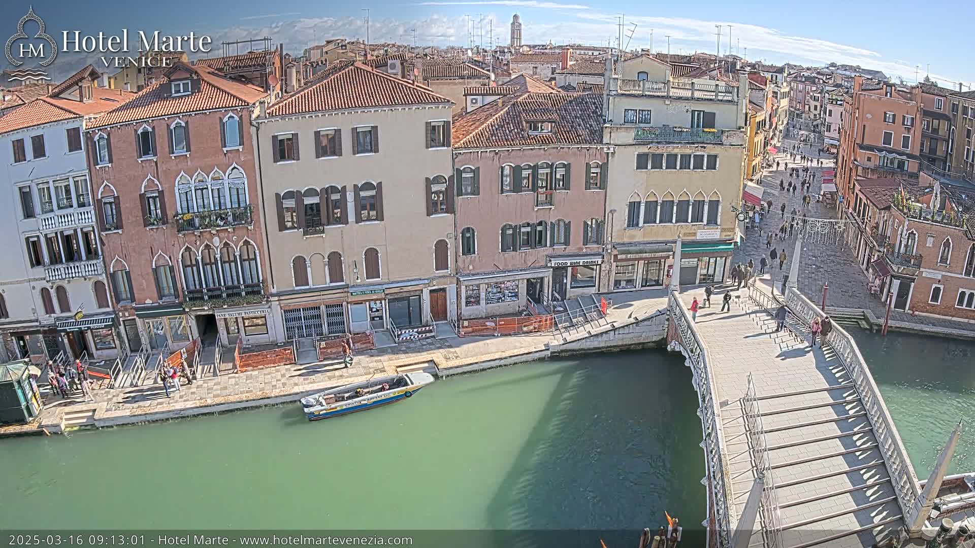 A sunny day in Venice shows a canal with a pedestrian bridge, lined with buildings and people walking across the bridge and along the canal.