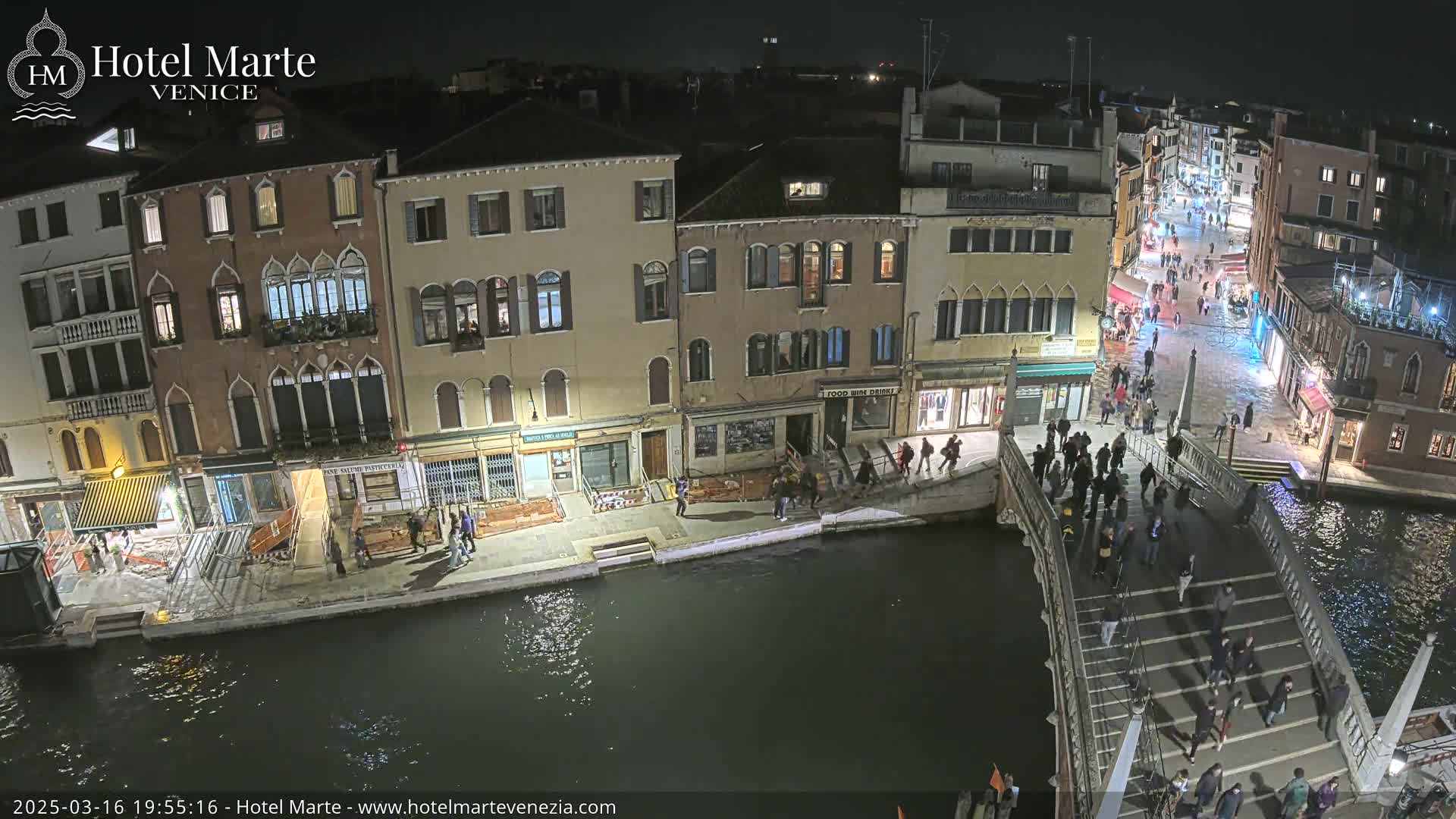 A nighttime aerial view shows a bridge in Venice with people walking across it and along the canals, under mostly clear skies.
