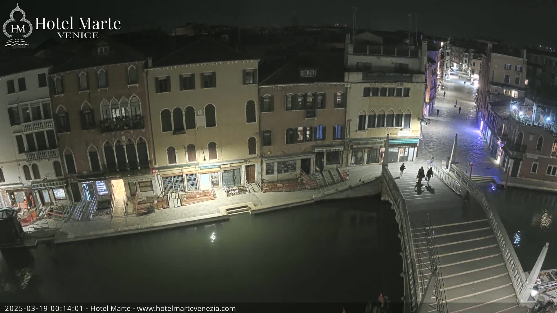 A nighttime view of a canal in Venice, showing several buildings along the waterway, a bridge with people walking across it, and calm water.