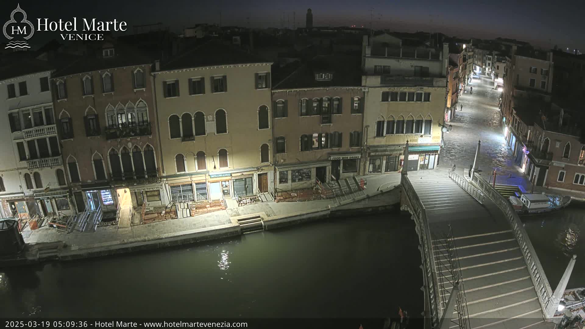 A nighttime view of a canal in Venice, showing several buildings lit up along the waterway, a bridge with steps leading down to the water, and a boat moored nearby.