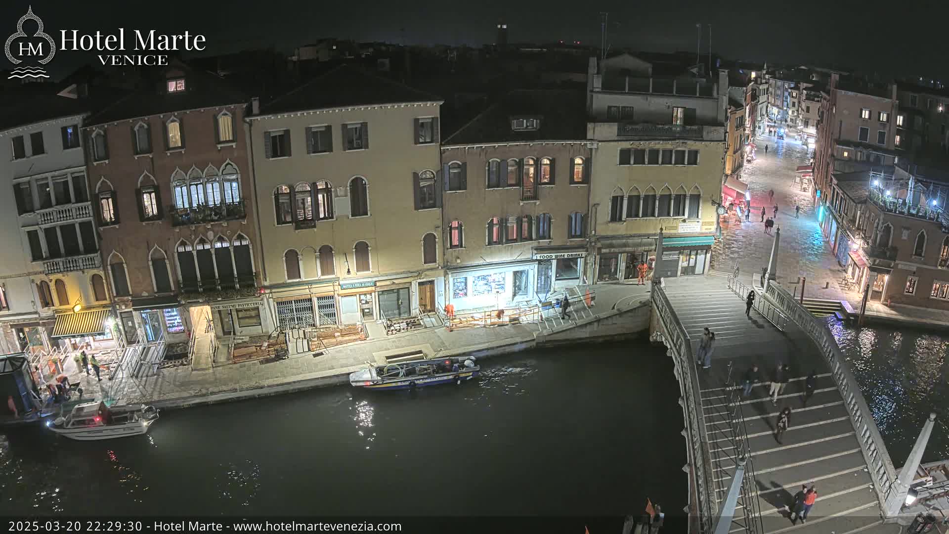 A nighttime view of a canal in Venice, showing several buildings, a bridge with pedestrians, and a boat on the water.