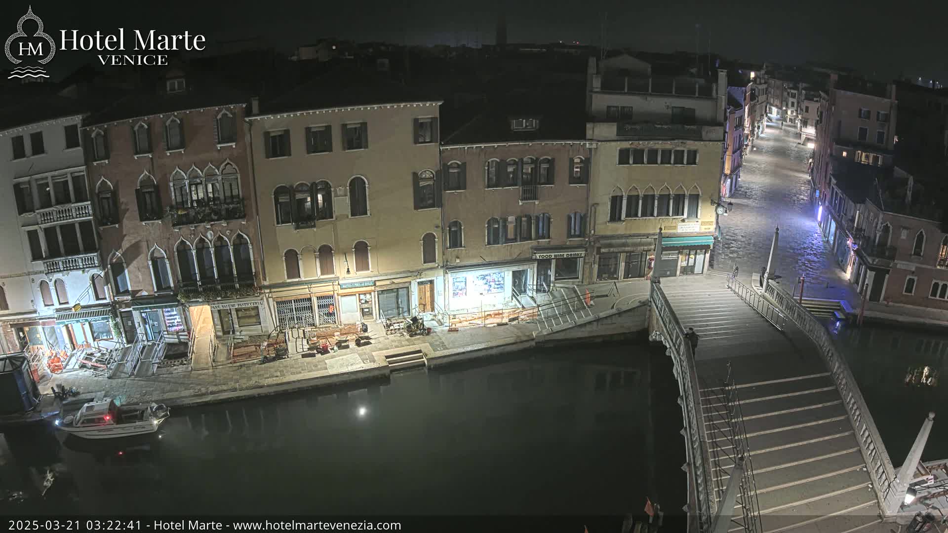 A nighttime view of a canal in Venice, showing several buildings, a bridge, and a small boat, with the canal partially flooded.