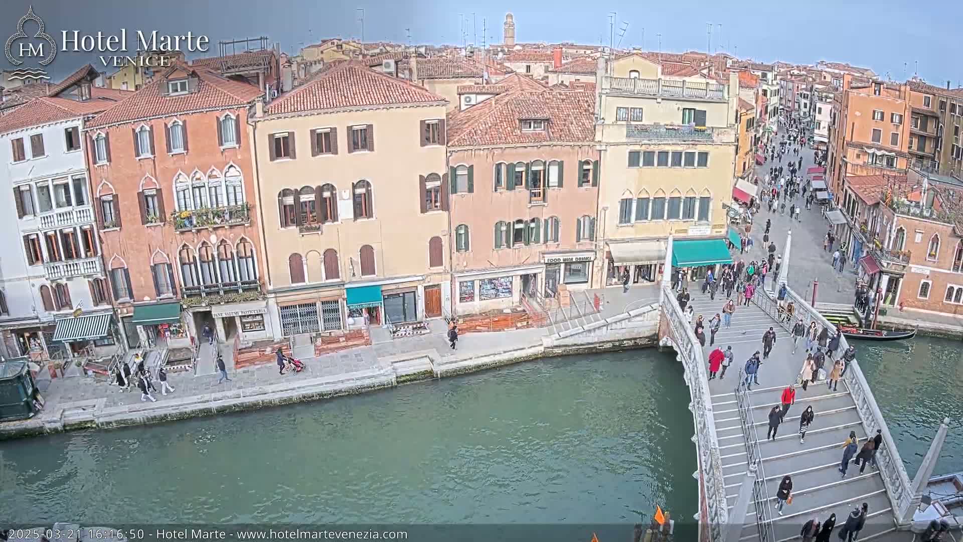 A sunny day in Venice shows a canal with pedestrians walking across a bridge and along the waterfront, lined with buildings.