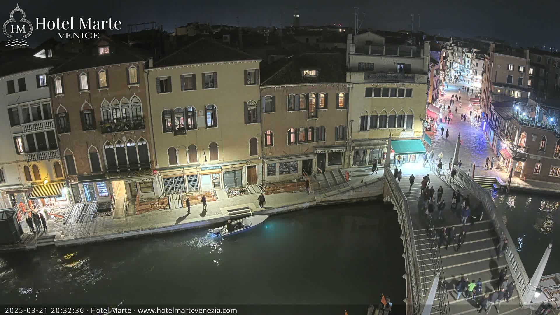 A nighttime view of a Venetian canal showing several buildings, pedestrians crossing a bridge, and a small boat on the water.