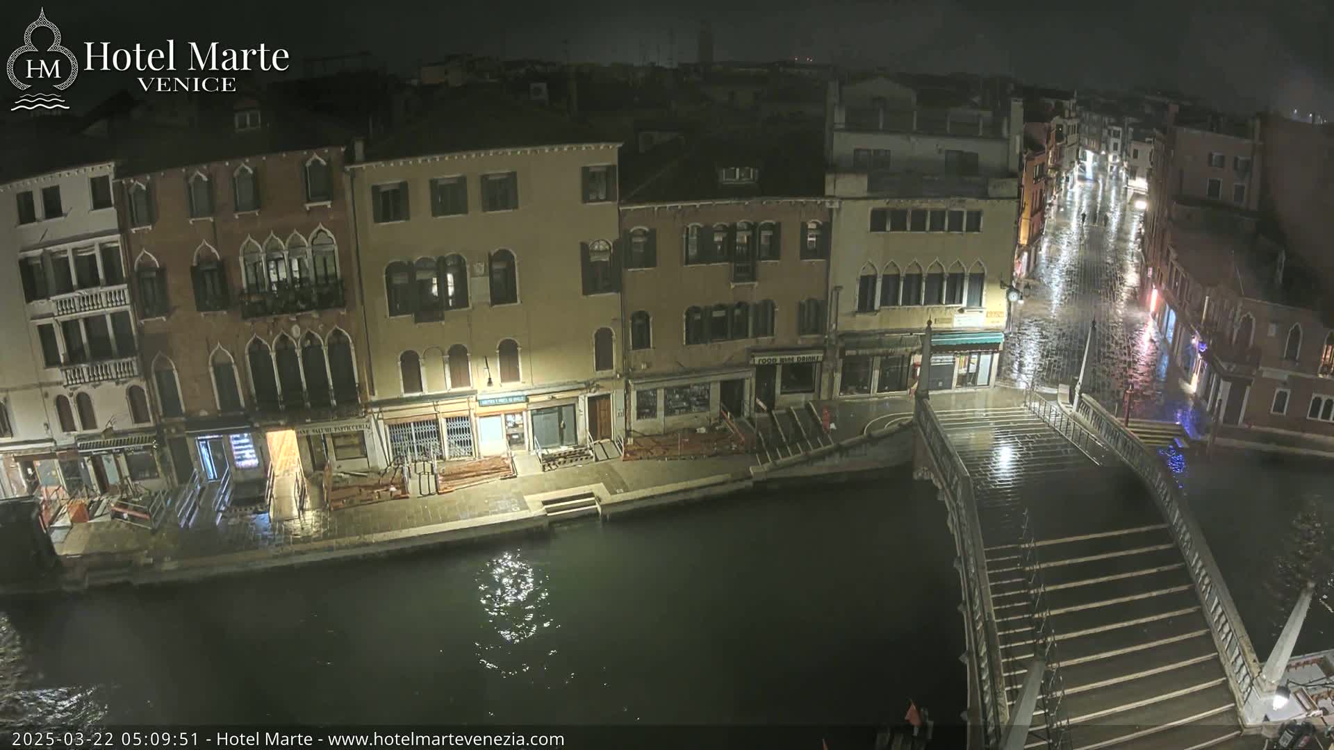 A nighttime view of a canal in Venice, showing several buildings along the waterfront, a bridge with steps flooded by rain, and wet cobblestone streets.
