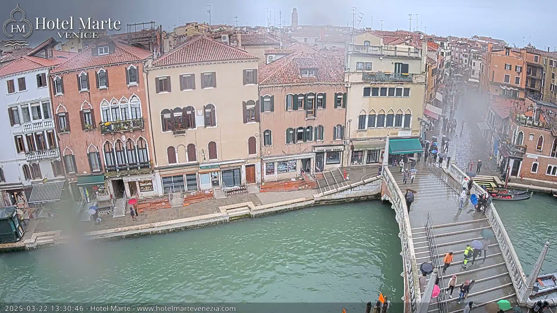 A rain-soaked canal in Venice, with people walking across a bridge and gondolas moored alongside colorful buildings.