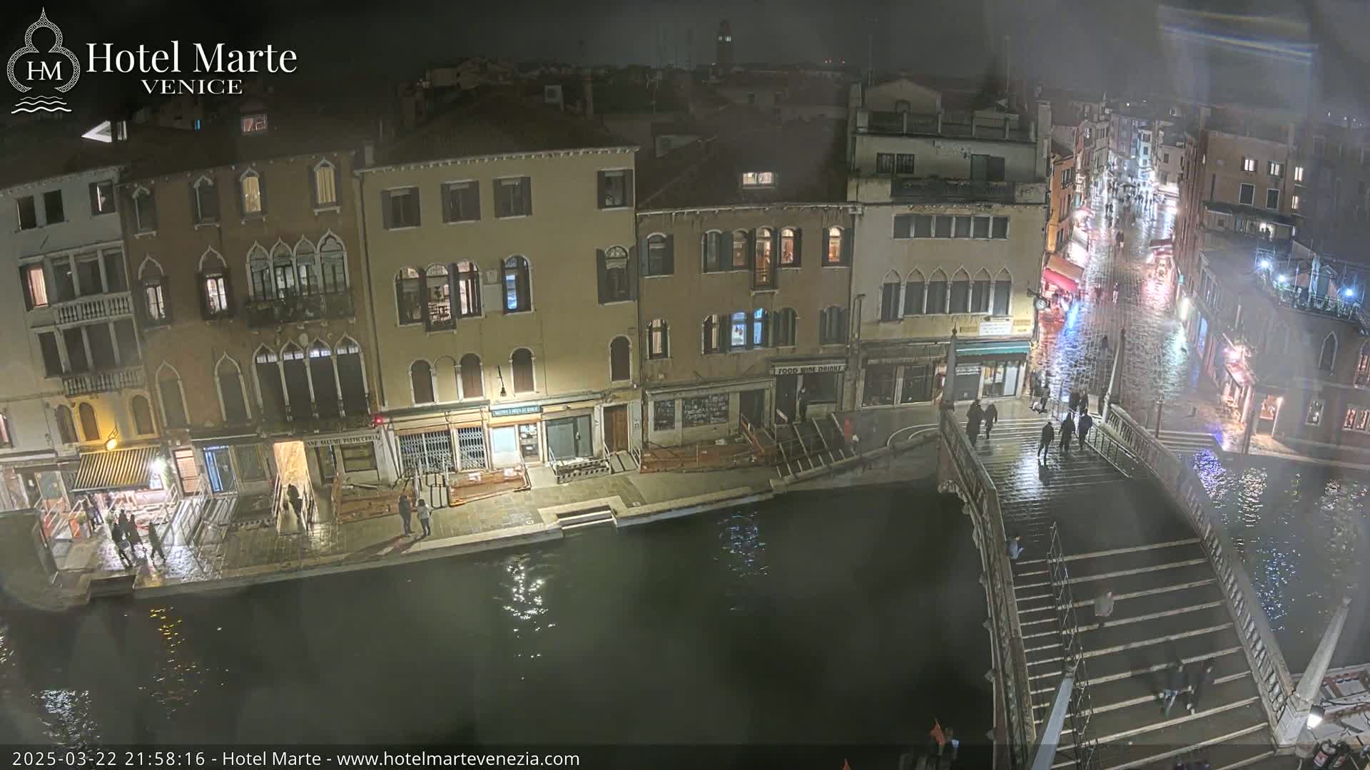 A nighttime view of a canal in Venice, showing several buildings, a bridge with people walking on it, and water at a high level.
