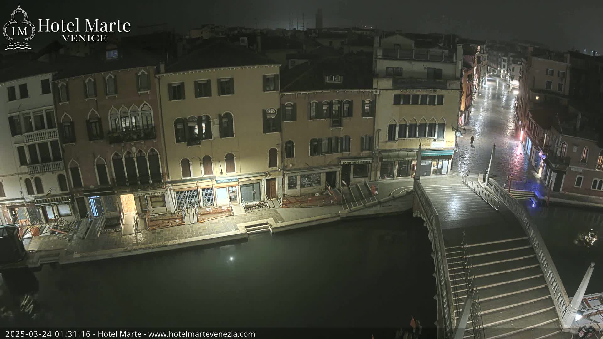 At night, under a dark sky, a canal in Venice shows several buildings lining its banks, a bridge crossing the water, and a wet street visible beyond the bridge.