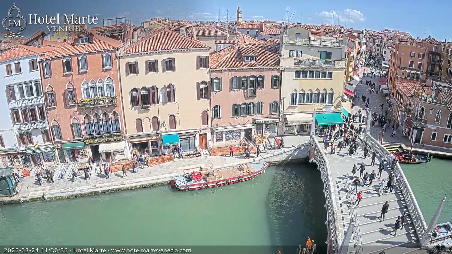 A sunny day in Venice shows a canal with a bridge crowded with pedestrians, boats, and buildings lining the waterway.