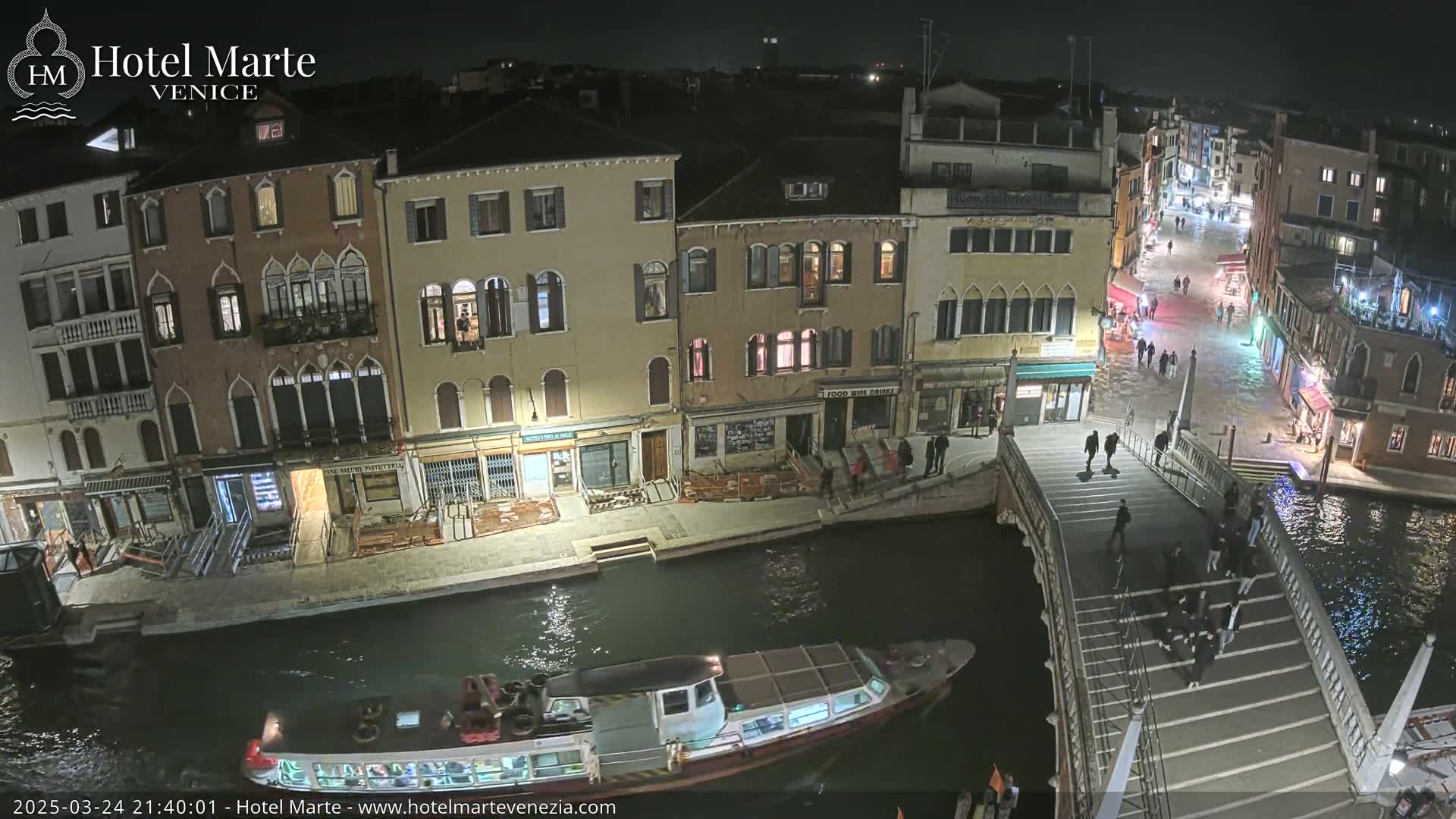 A nighttime aerial view of a Venetian canal shows several buildings lining the waterway, a bridge with pedestrians, and a boat moored nearby.