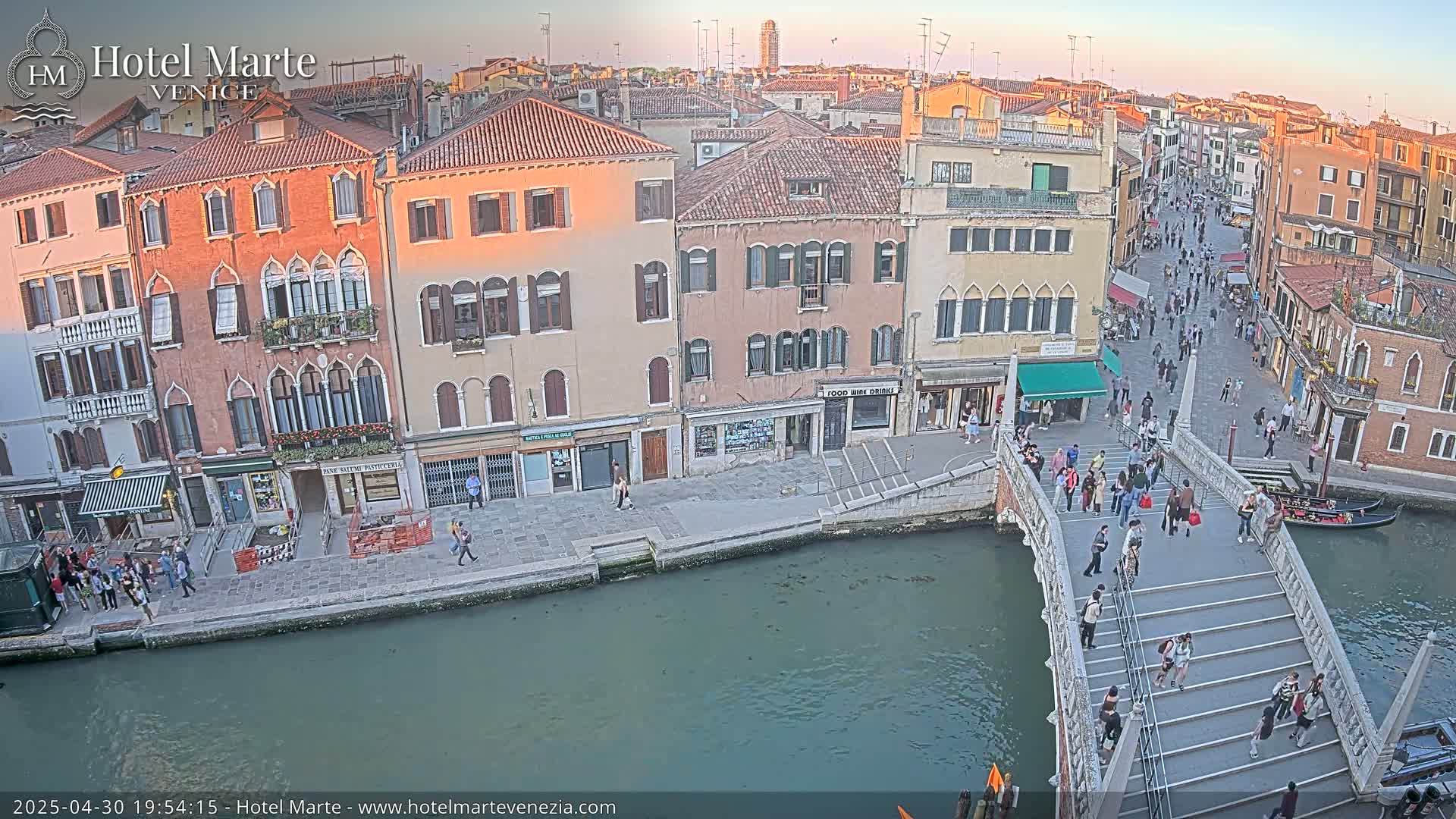 A Venetian canal at sunset, with pedestrians walking on a bridge and along the canalsides past colorful buildings.
