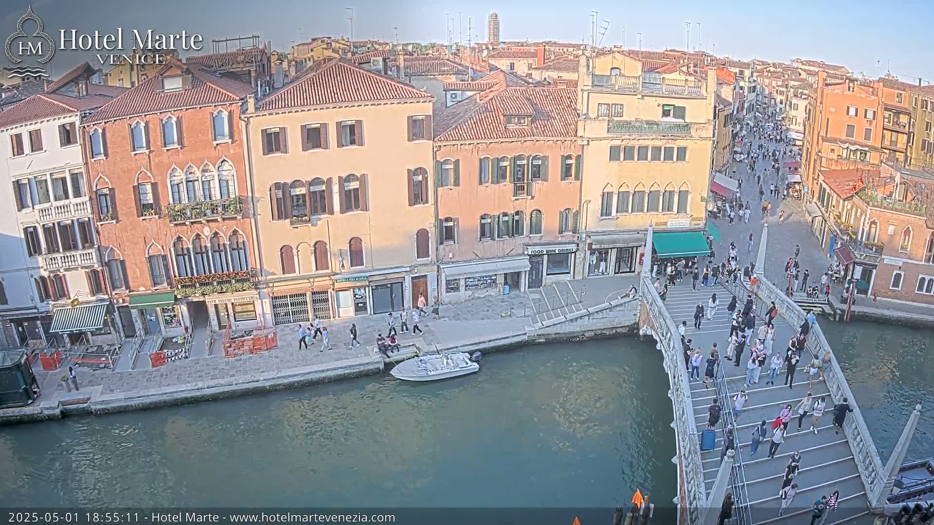 A canal in Venice, under a clear sky, is filled with boats and people walking across a bridge lined with buildings.
