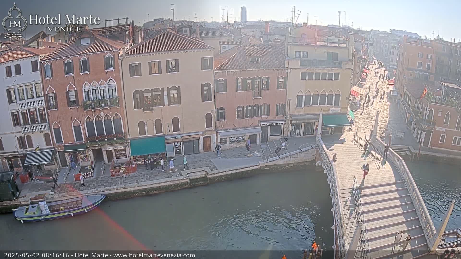 A sunny view of a canal in Venice, showing several buildings, a bridge with pedestrians, and a boat.