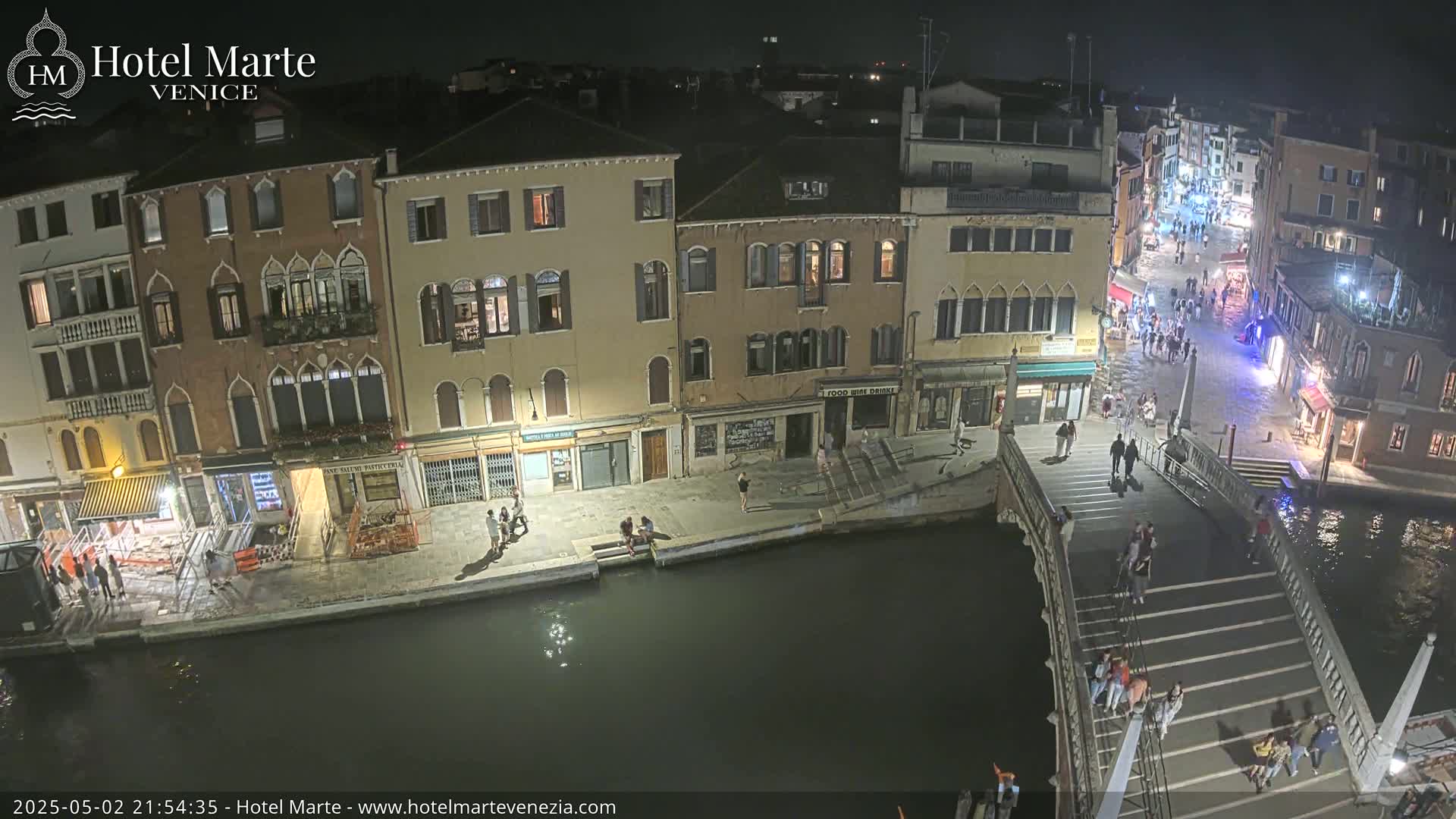 At night, a canal-side view shows illuminated buildings, pedestrians on a bridge and along a waterfront, and a dark canal.